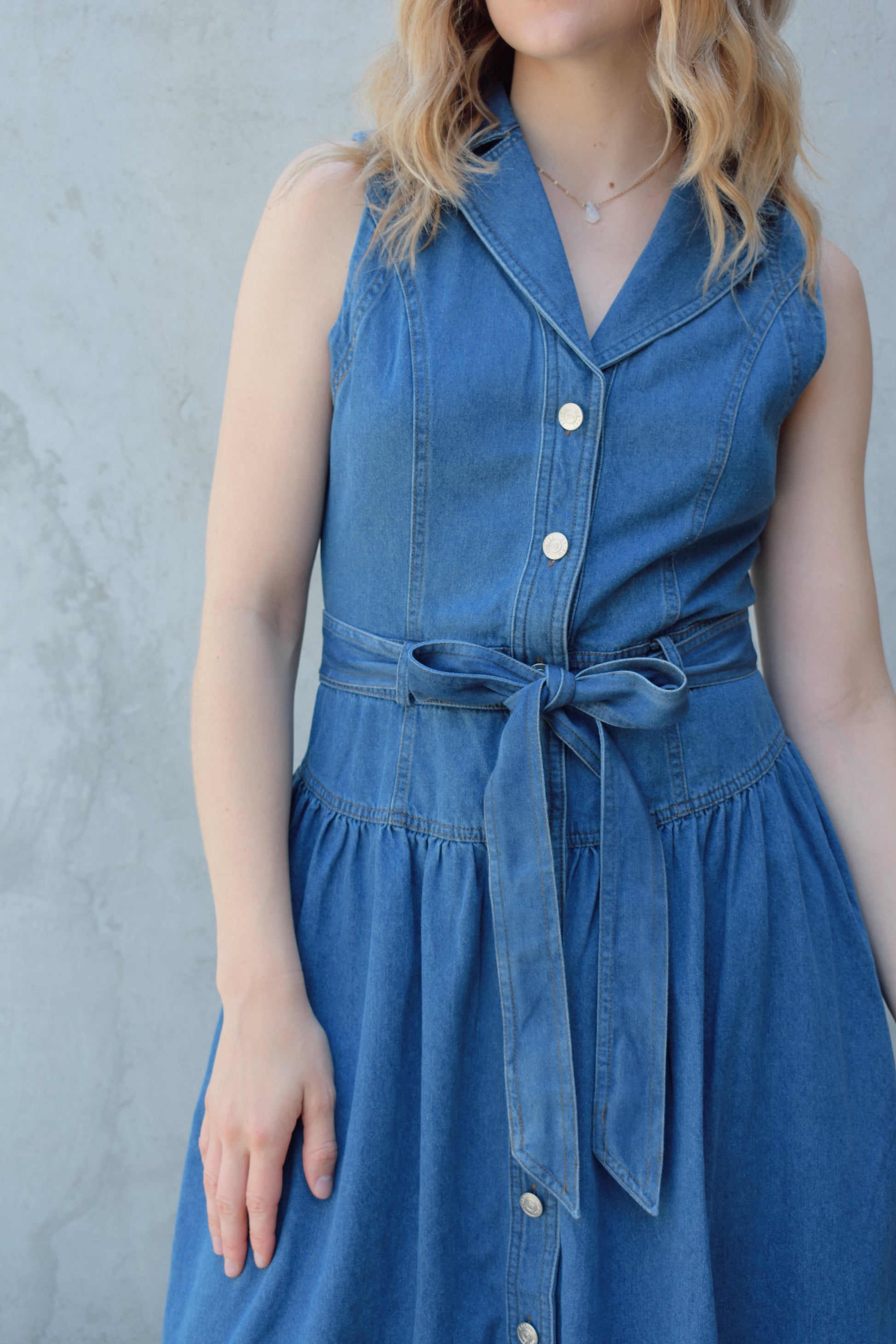 sleeveless medium wash denim midi dress with notched lapel collar, buttons down the entire front, drop waist, beltloops with denim tie at waist, has pockets