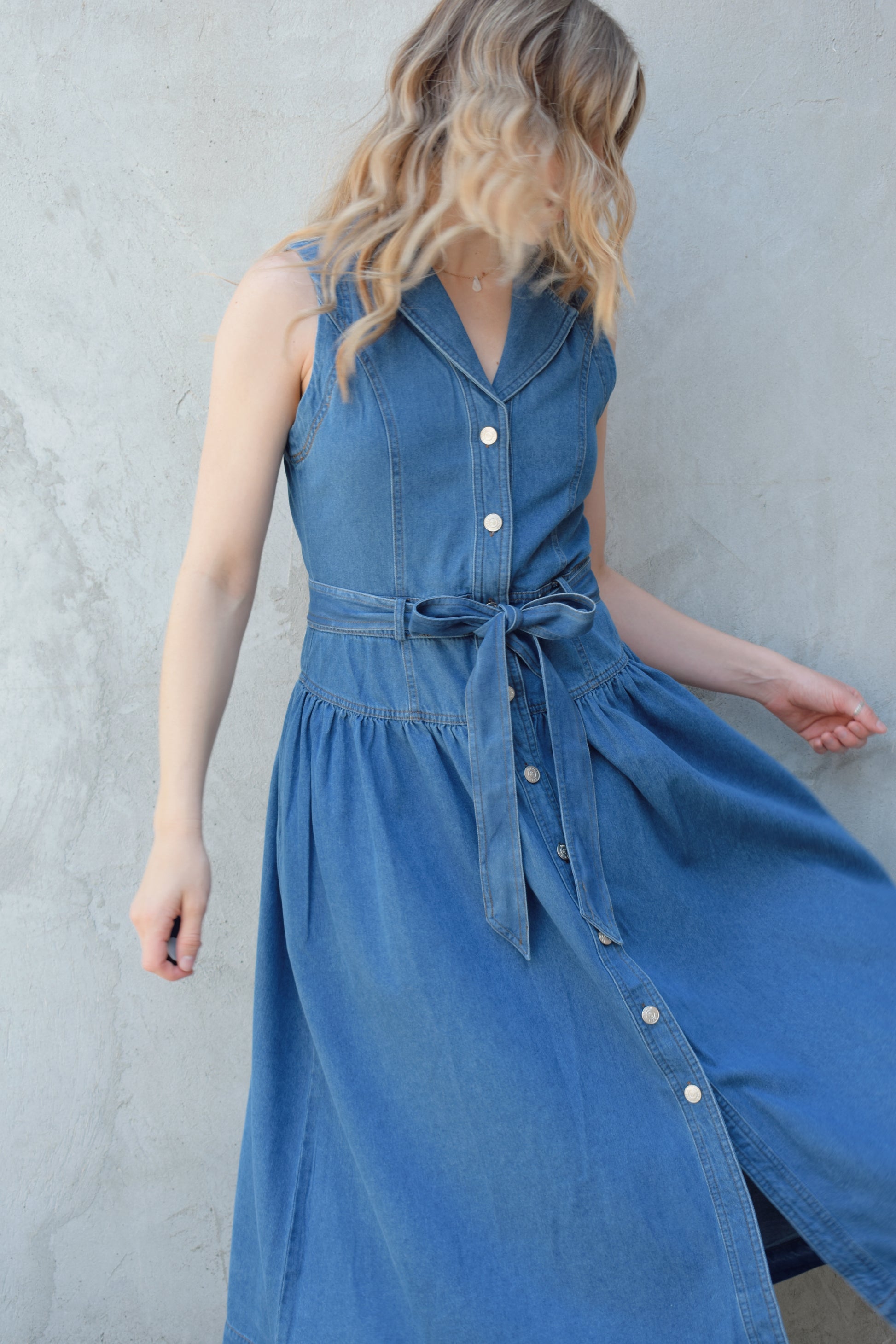 sleeveless medium wash denim midi dress with notched lapel collar, buttons down the entire front, drop waist, beltloops with denim tie at waist, has pockets