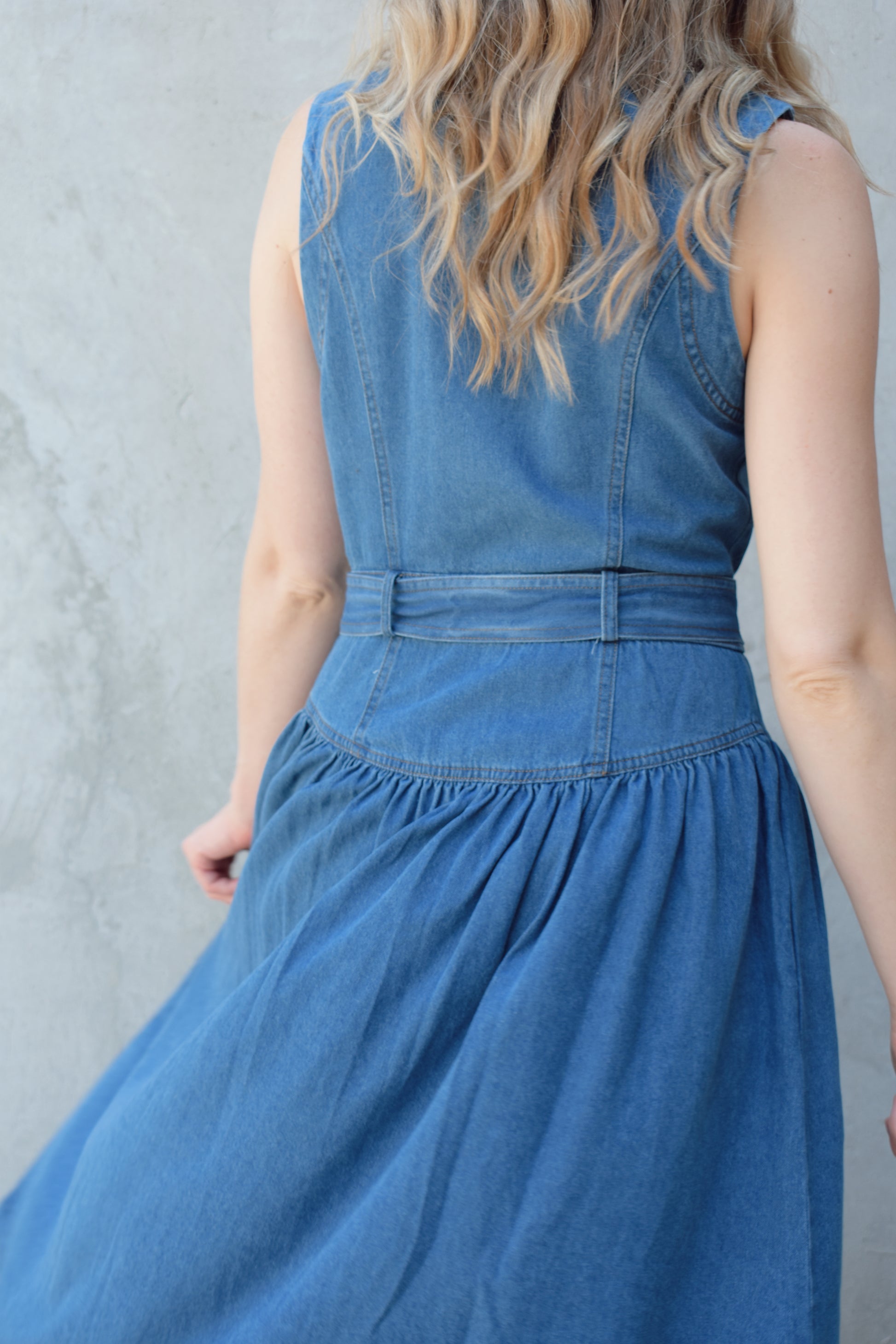 sleeveless medium wash denim midi dress with notched lapel collar, buttons down the entire front, drop waist, beltloops with denim tie at waist, has pockets