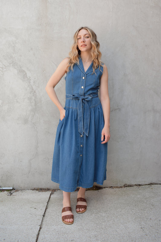 sleeveless medium wash denim midi dress with notched lapel collar, buttons down the entire front, drop waist, beltloops with denim tie at waist, has pockets