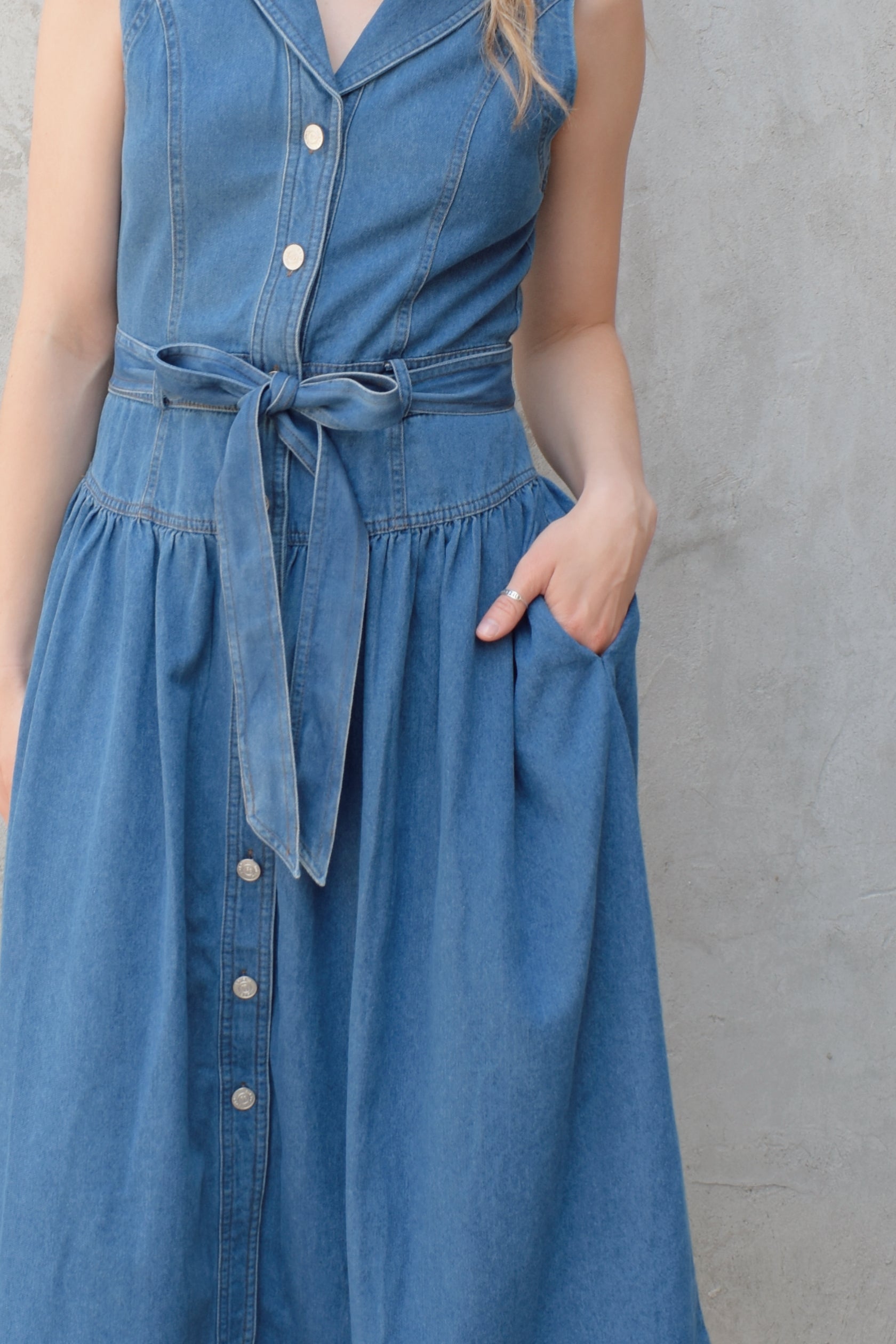 sleeveless medium wash denim midi dress with notched lapel collar, buttons down the entire front, drop waist, beltloops with denim tie at waist, has pockets