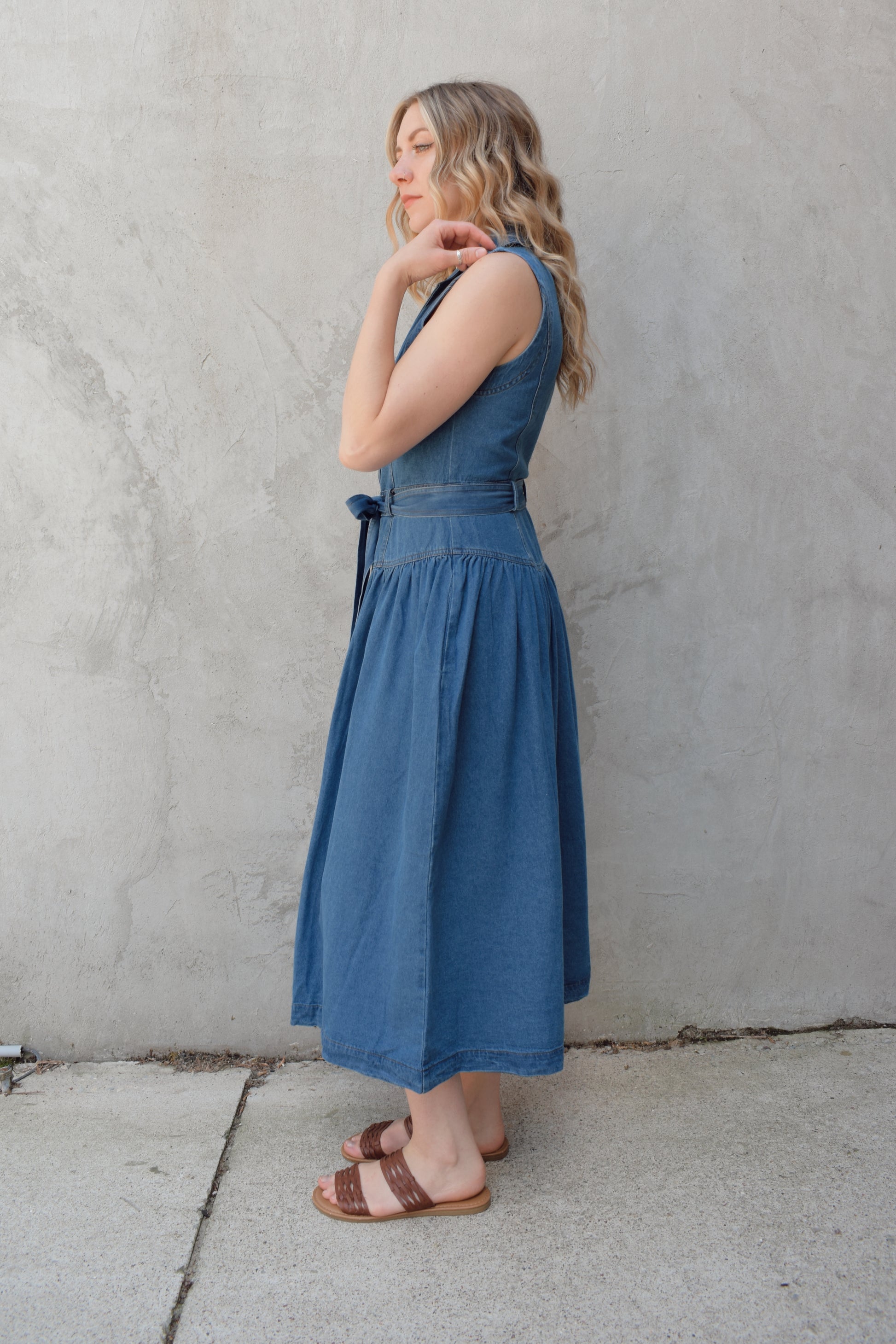 sleeveless medium wash denim midi dress with notched lapel collar, buttons down the entire front, drop waist, beltloops with denim tie at waist, has pockets
