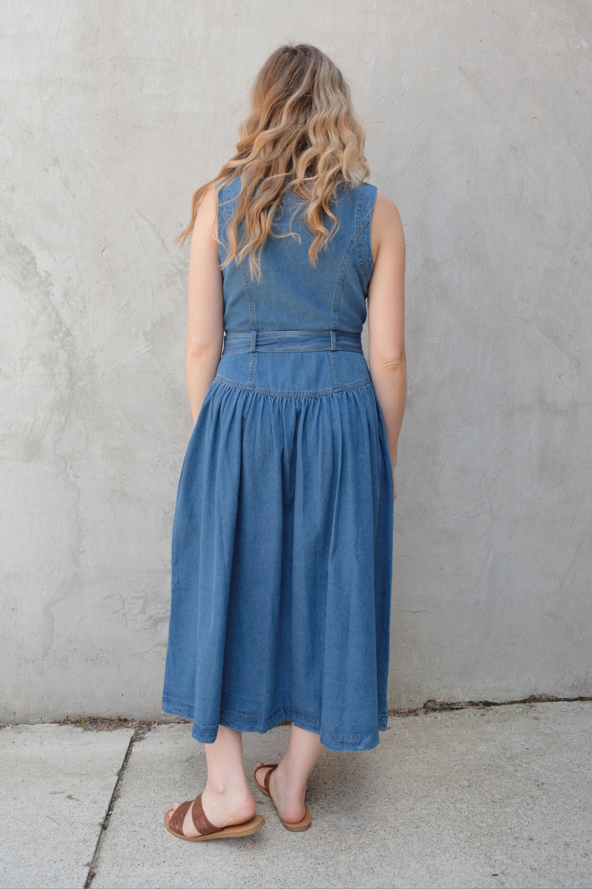 sleeveless medium wash denim midi dress with notched lapel collar, buttons down the entire front, drop waist, beltloops with denim tie at waist, has pockets