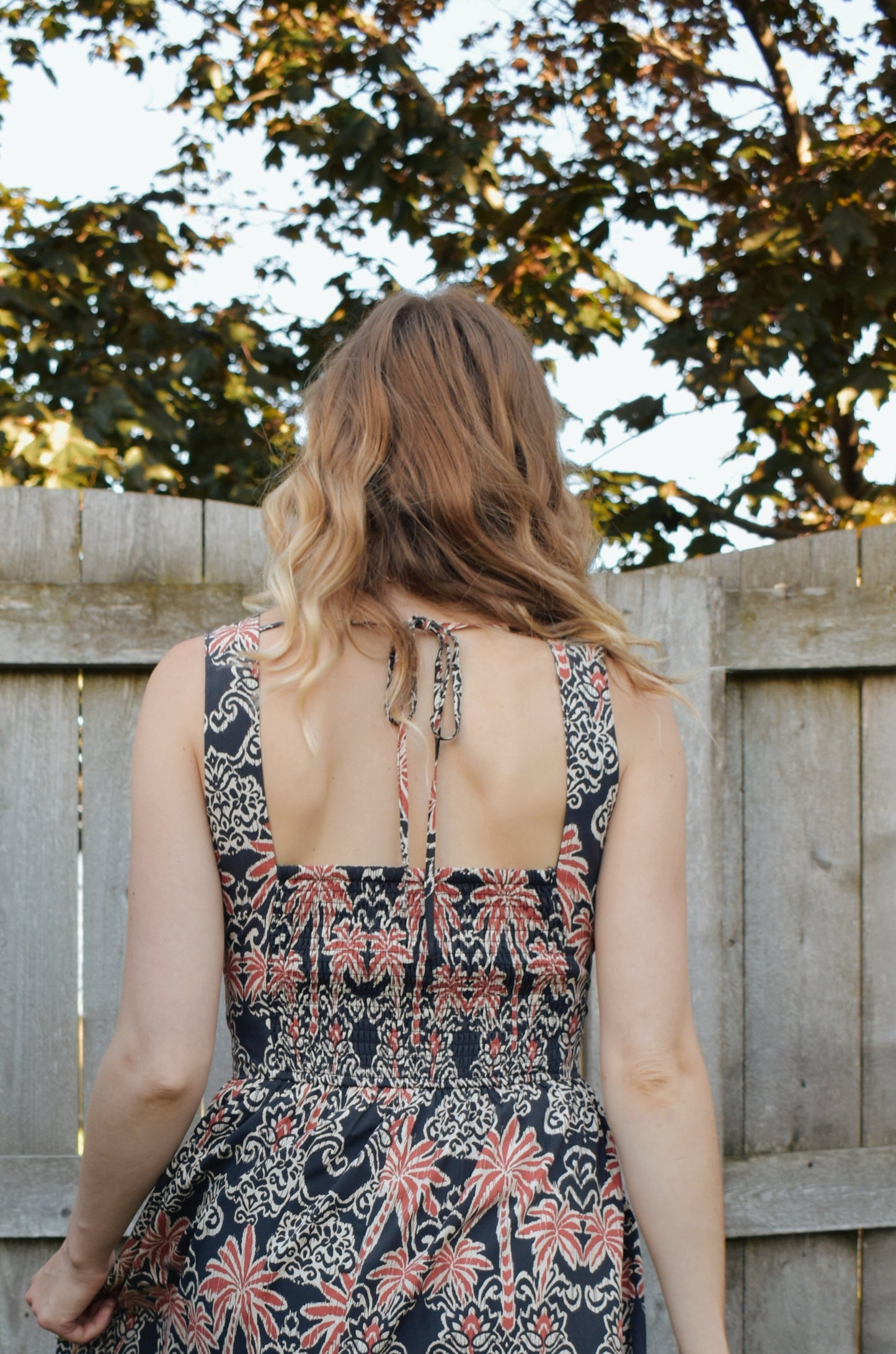 v neck midi dress with oval cut outs at waistline with circle loop in the center. square neck on back with tie across the top. sleeveless with thick tank straps. dark blue background with white and light red summer print. smocked on back waistline for stretch.