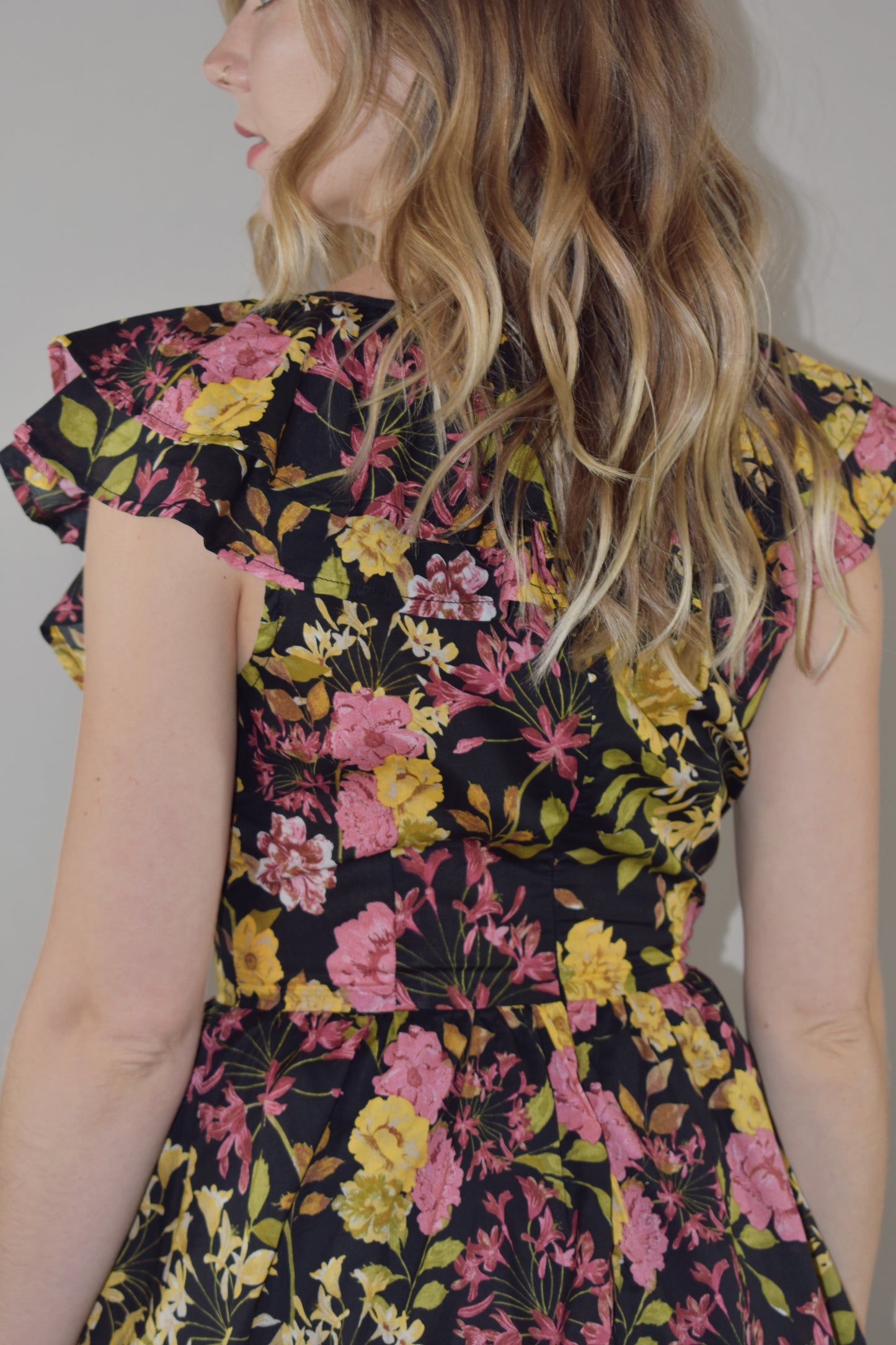 midi tiered floral dress with black background and pink, yellow, green and neutral tones in the floral pattern. v neck with cap flutter sleeves that are also layered. fitted bodice and invisible back zip enclosure. 