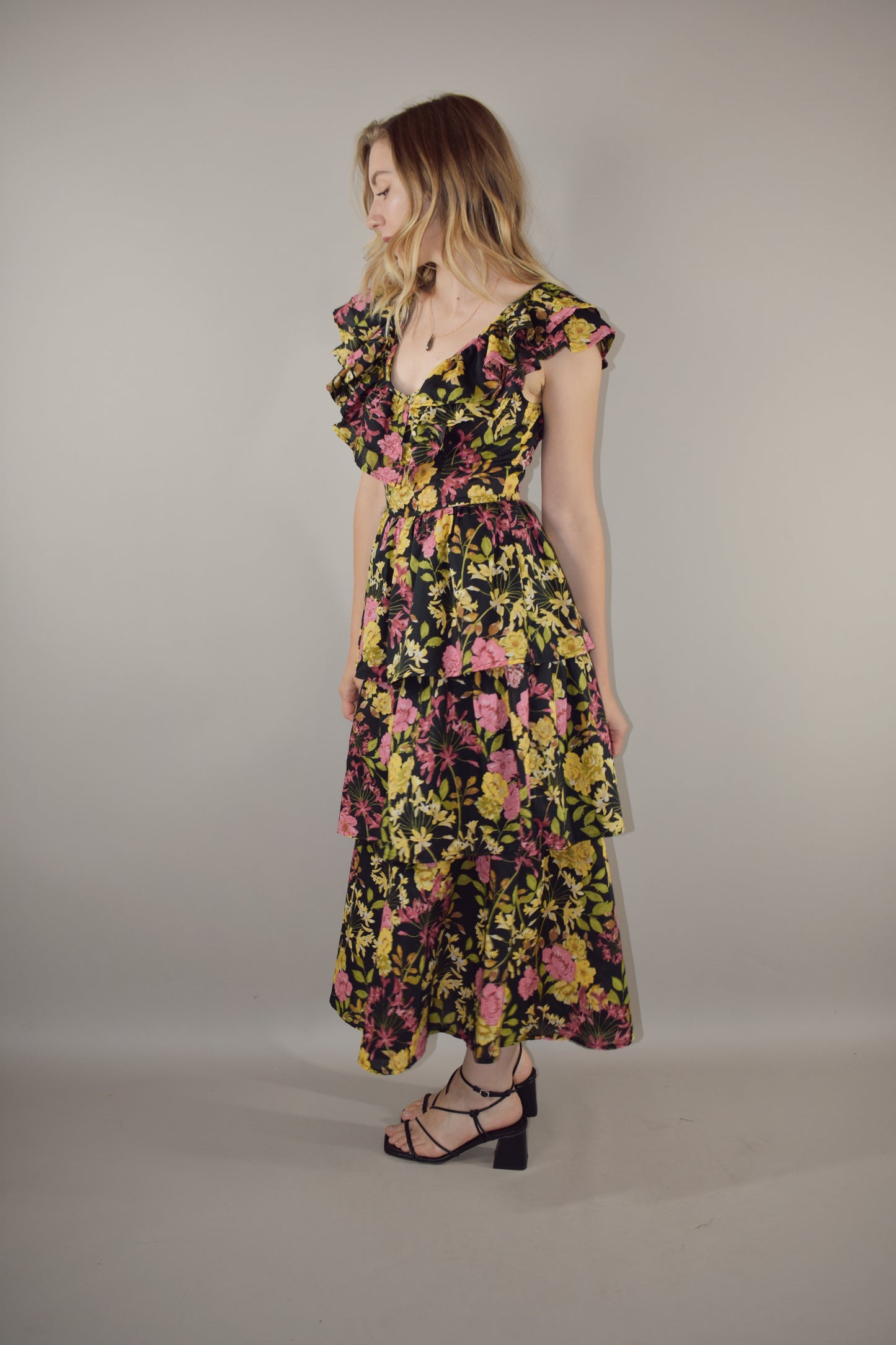 midi tiered floral dress with black background and pink, yellow, green and neutral tones in the floral pattern. v neck with cap flutter sleeves that are also layered. fitted bodice and invisible back zip enclosure. 