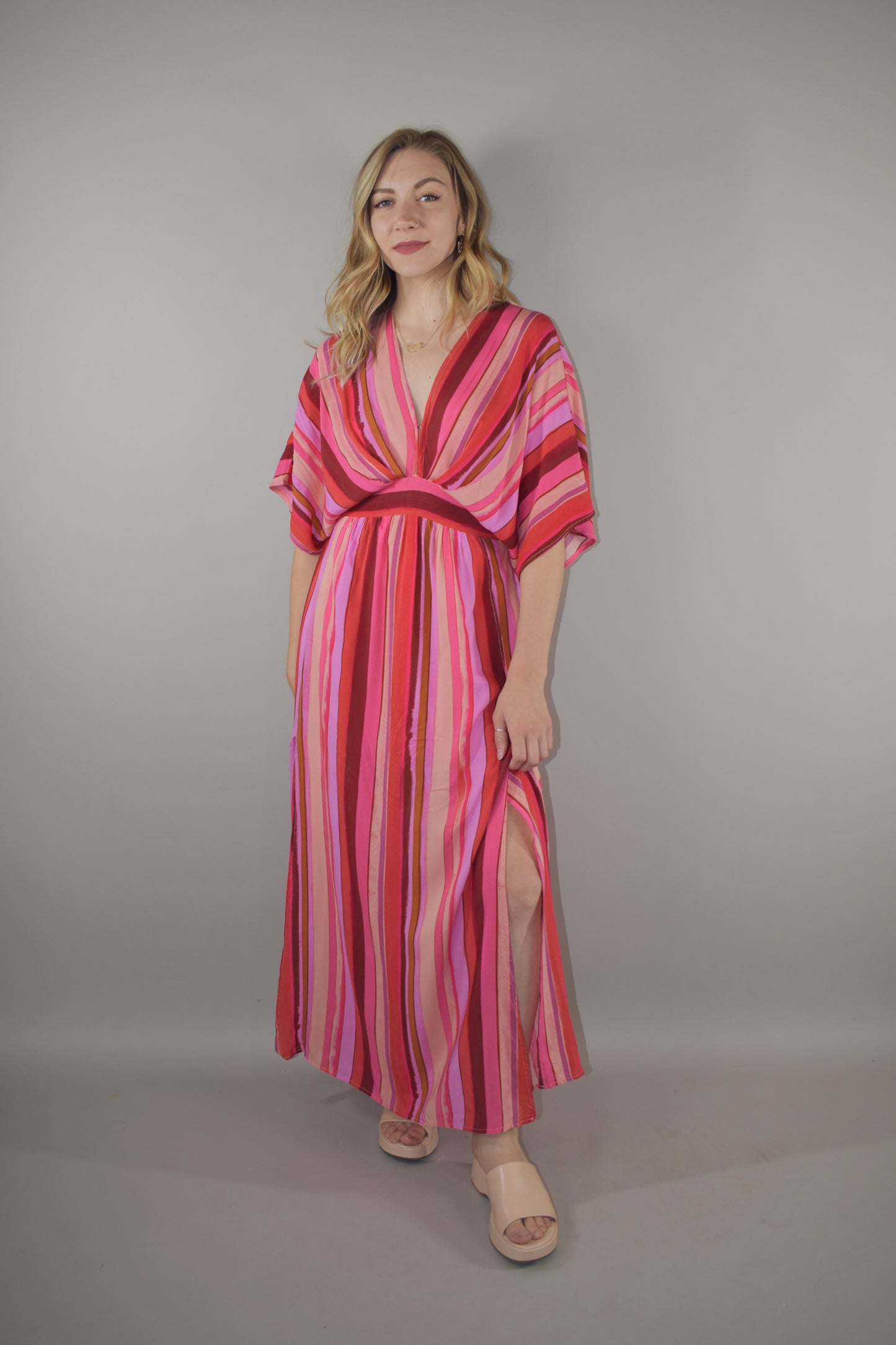 tones of red vertical striped maxi dress with flowy lightweight fabric, lined with a synched stretchy waistband and deep v neck with a hook and eye enclosure and dolman sleeves that hit at the elbow. side slits that are semi hidden unless walking. button enclosure at back neckline