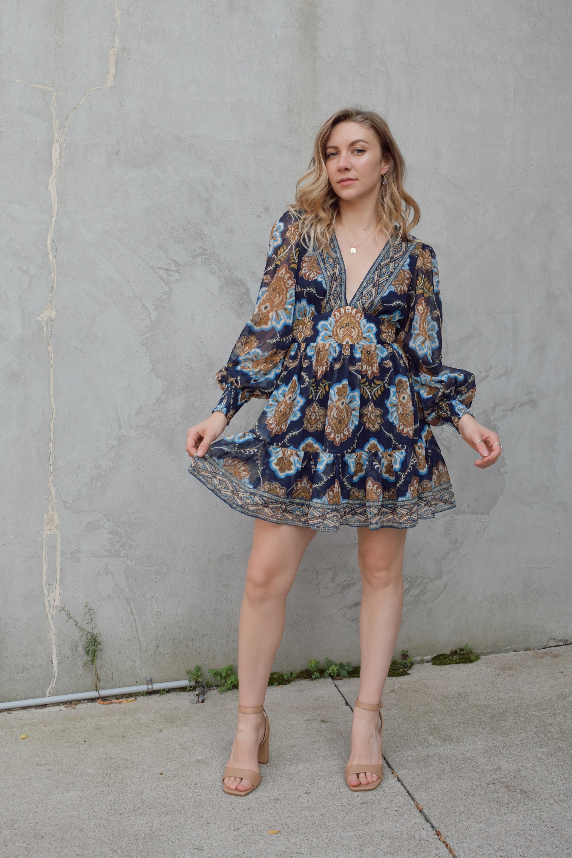 dark blue mini dress with gold, white, and light blue boho pattern. deep v neckline and waist yoke with long sleeves sinched at cuff. back zip enclosure. full skirt. chiffon fabric making sleeves look sheer.