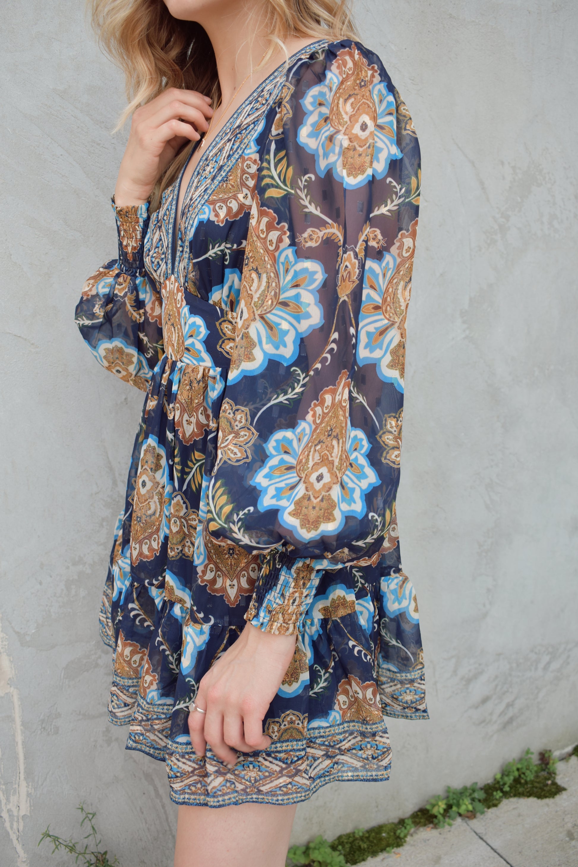 dark blue mini dress with gold, white, and light blue boho pattern. deep v neckline and waist yoke with long sleeves sinched at cuff. back zip enclosure. full skirt. chiffon fabric making sleeves look sheer.